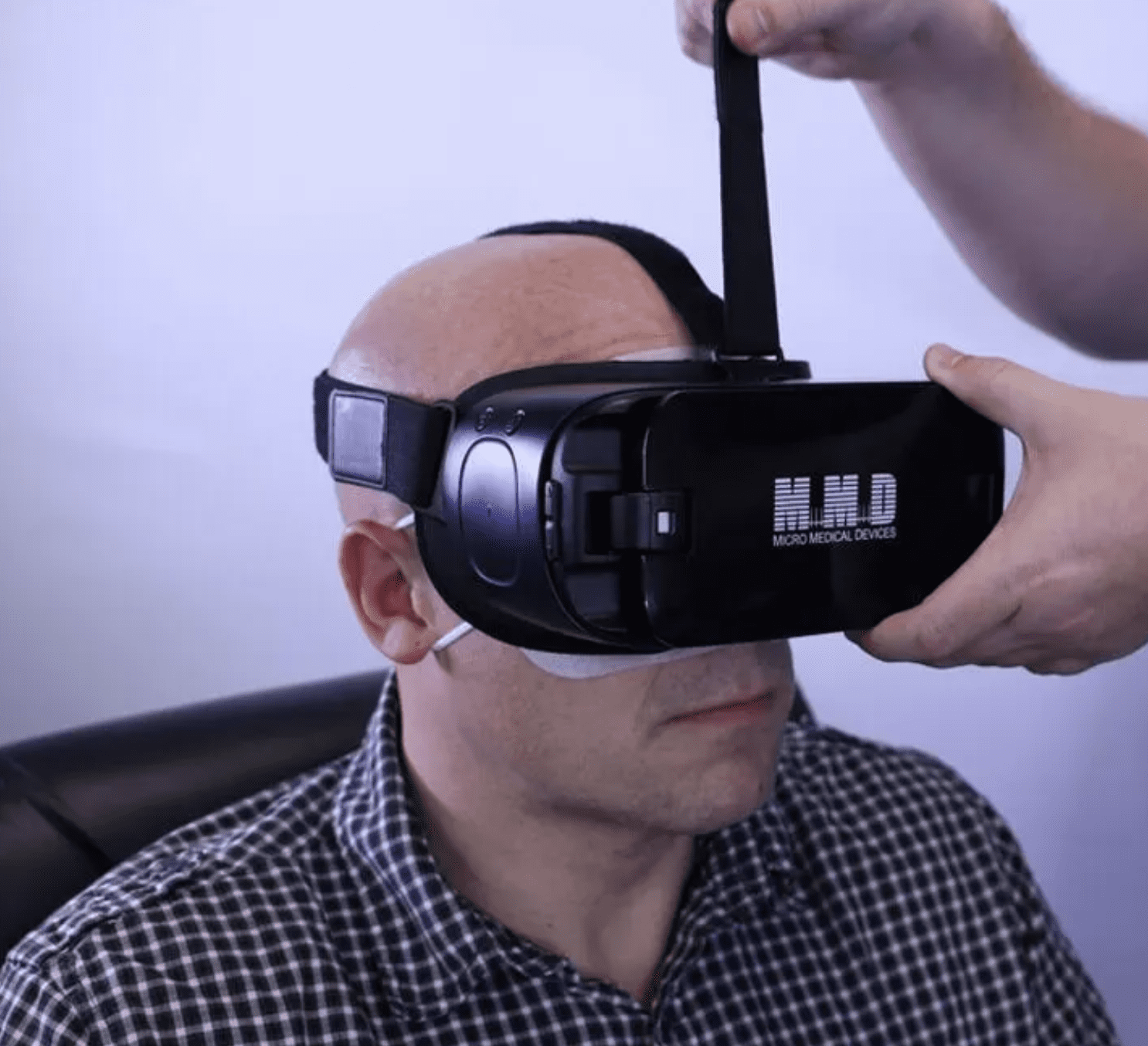 A person wearing a virtual reality headset labeled HMD Micro Medical Devices is being assisted by another person adjusting a strap on their head. The person is seated and wearing a checkered shirt.